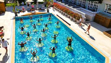 A swimming pool with people exercising in the water, surrounded by outdoor seating and a building with a balcony.