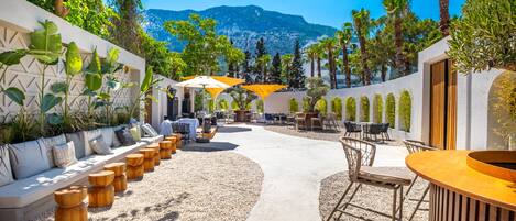 An outdoor patio area with seating, tables, and umbrellas, surrounded by greenery and a mountain view.