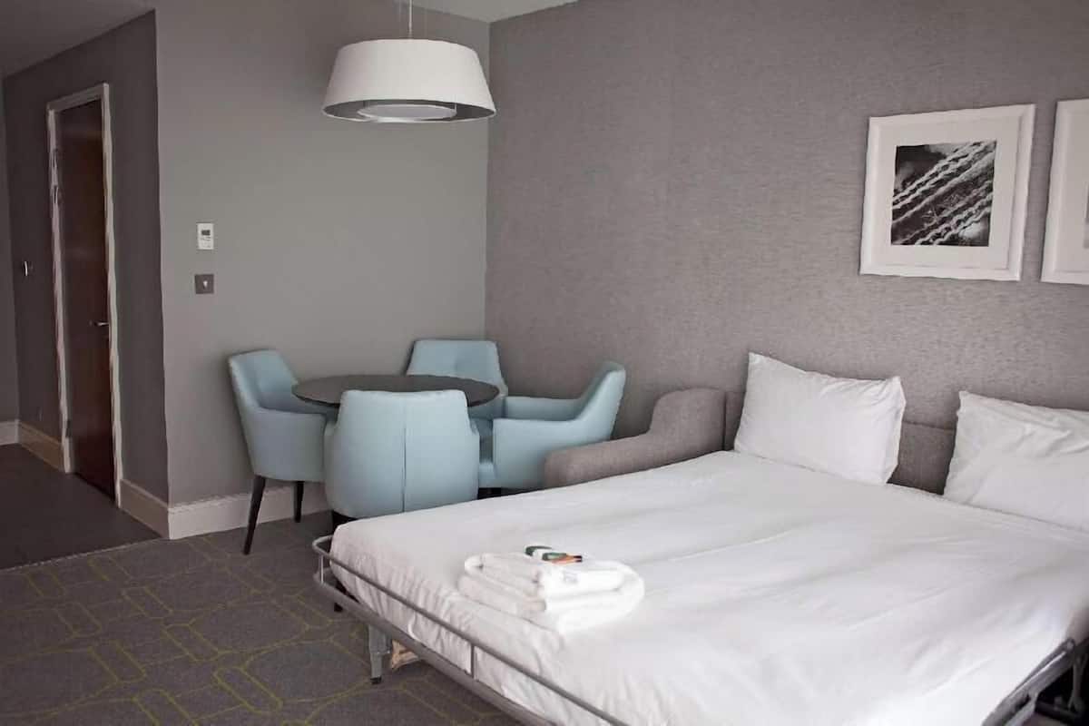 A hotel room with a bed, a desk, two chairs, a lamp, and two framed pictures on the wall.