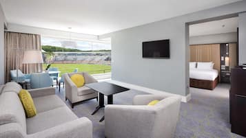 A hotel room with a seating area, a bed, a TV, and a view of a stadium.
