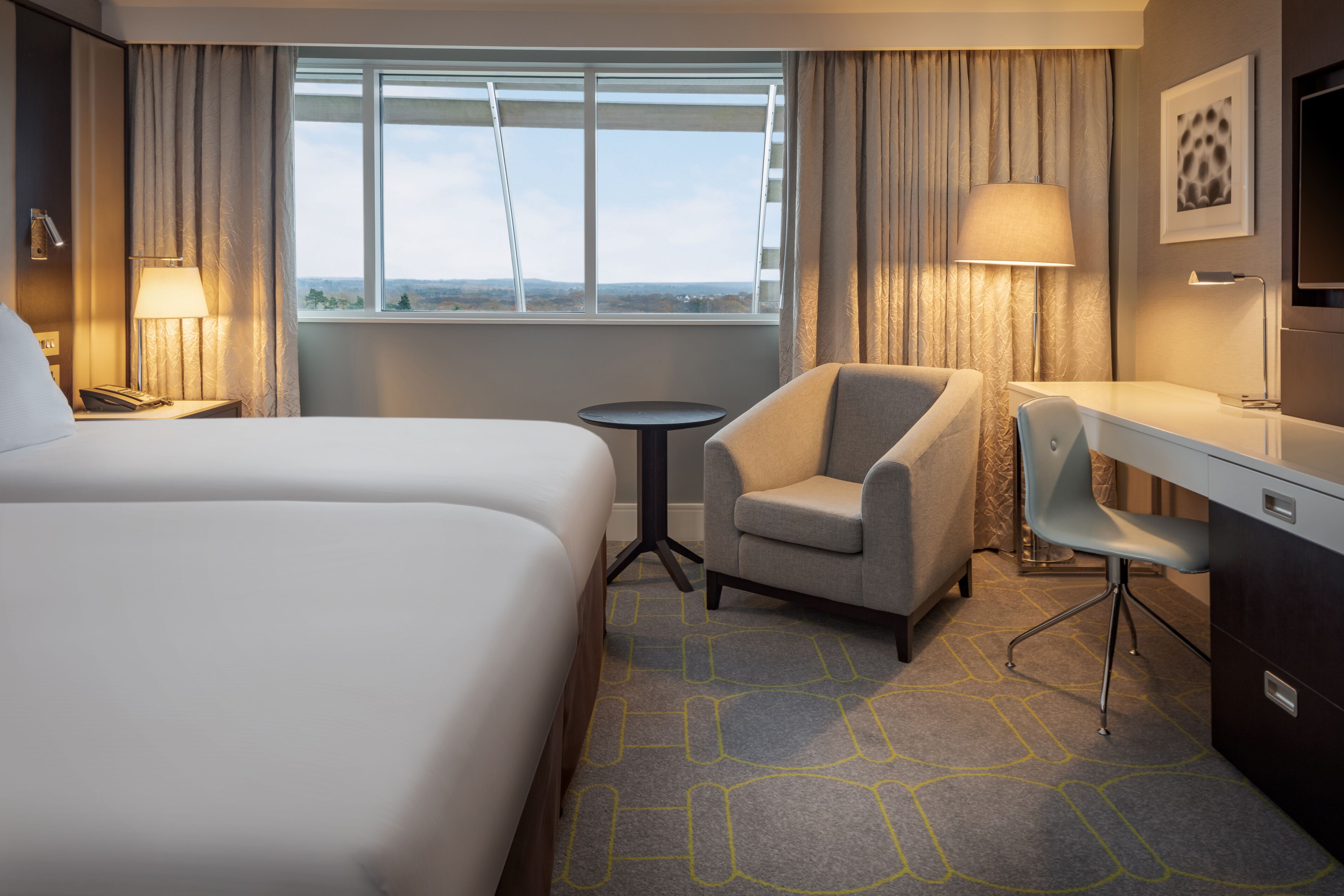 A hotel room with a large bed, a desk, a chair, a small table, and a window with curtains.