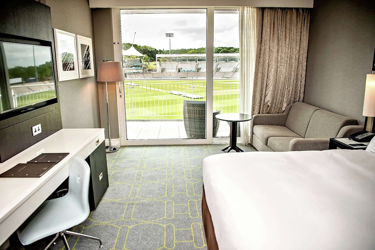 Een hotelkamer met een bed, een bureau, een stoel, een klein tafeltje, een televisie en uitzicht op een sportveld.