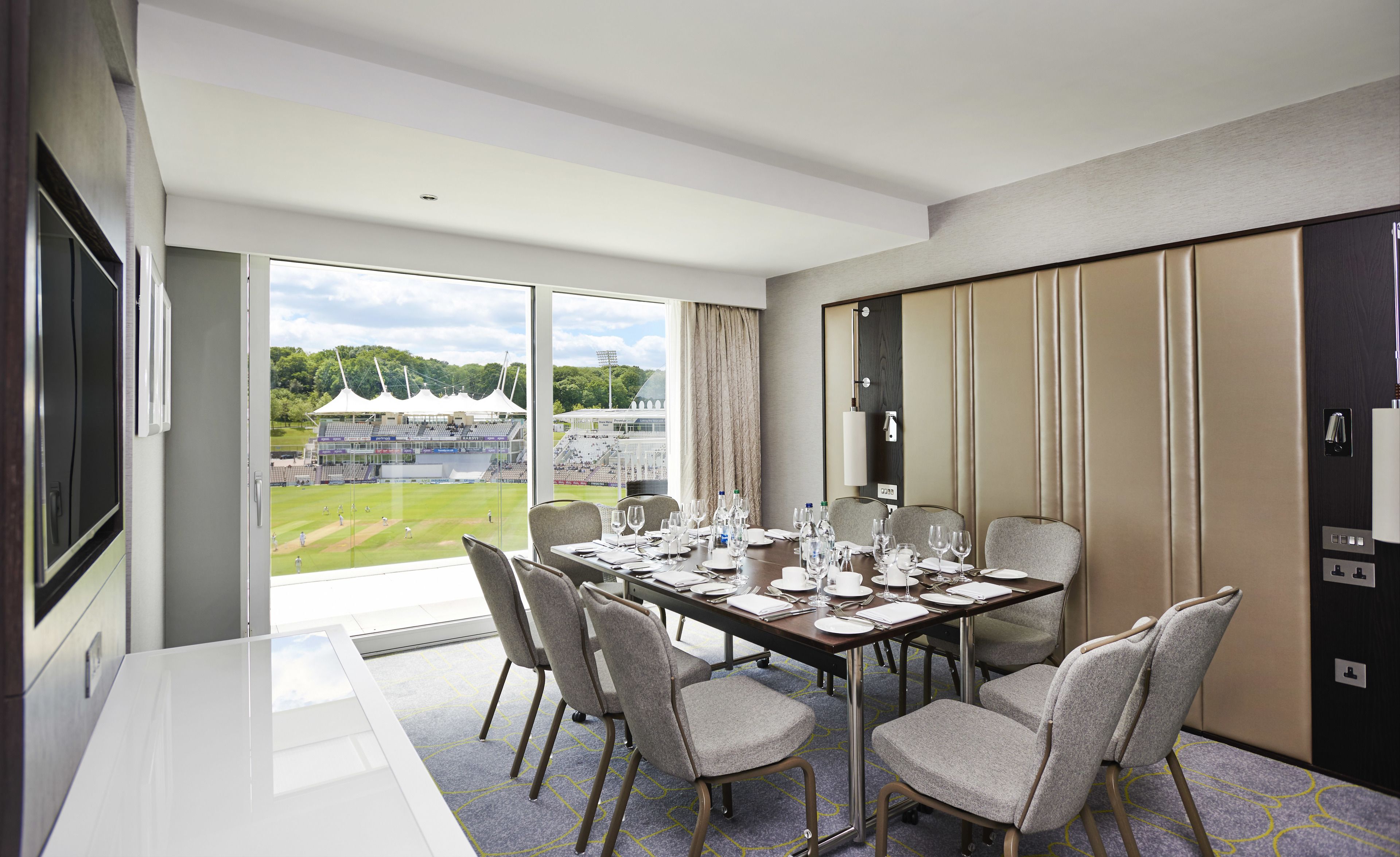A hotel room with a large window offering a view of a stadium. The room has a dining table set for six with chairs, a flat-screen TV mounted on the wall, and a door with a mirror and light switch.