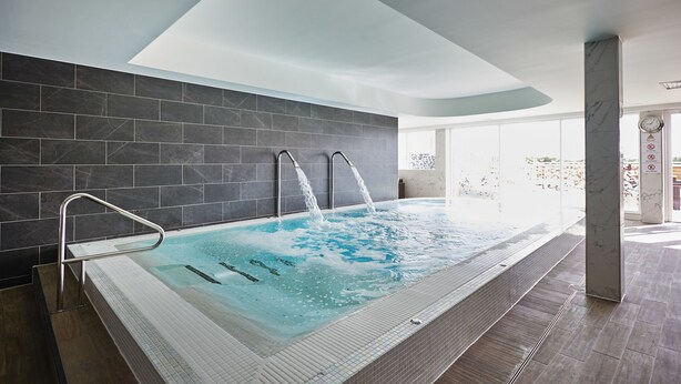 A modern indoor swimming pool with multiple water jets, a glass barrier, and a view of the outdoors.