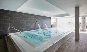 Een modern binnenzwembad met diverse waterstralen, een glazen afscheiding en uitzicht op het buitengebied.