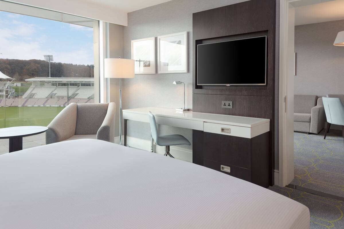 Hotelkamer met een groot bed, een bureau met stoel, een televisie en uitzicht op een sportveld.