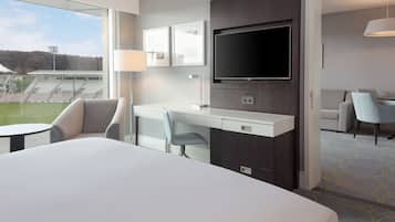 A hotel room with a large bed, a desk with a chair, a television, and a view of a sports field.