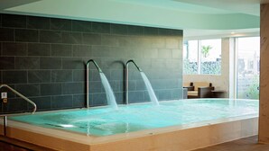 A large indoor swimming pool with two water jets, a tiled wall, and a seating area with a view of the outdoors.