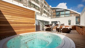 A cruise ship's outdoor swimming pool with wooden deck and lounge seating, surrounded by a modern building with a "Hilton" sign.