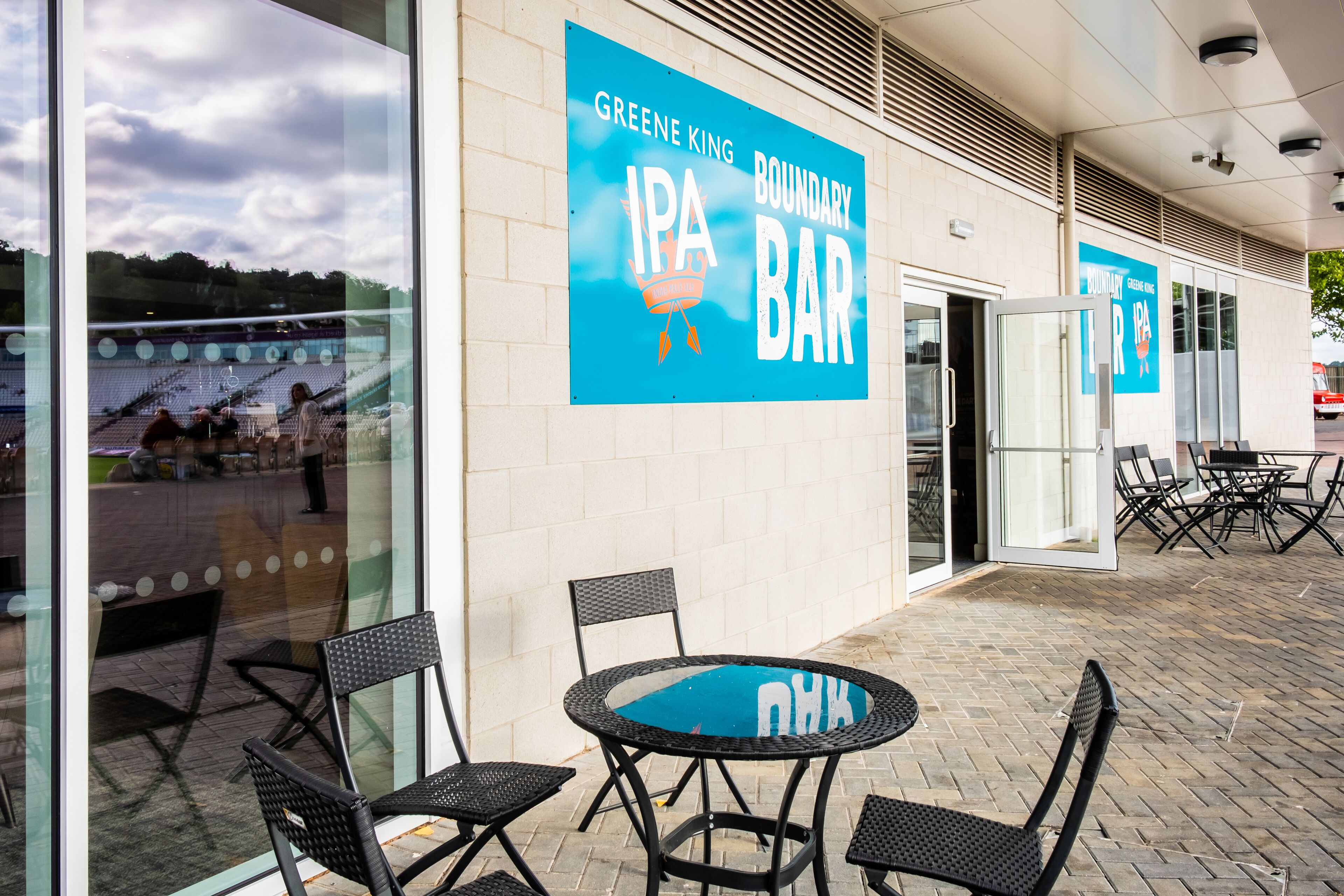 A patio area with a round table and four chairs, a glass wall reflecting a stadium, and a sign for Greene King Boundary IPA Bar.