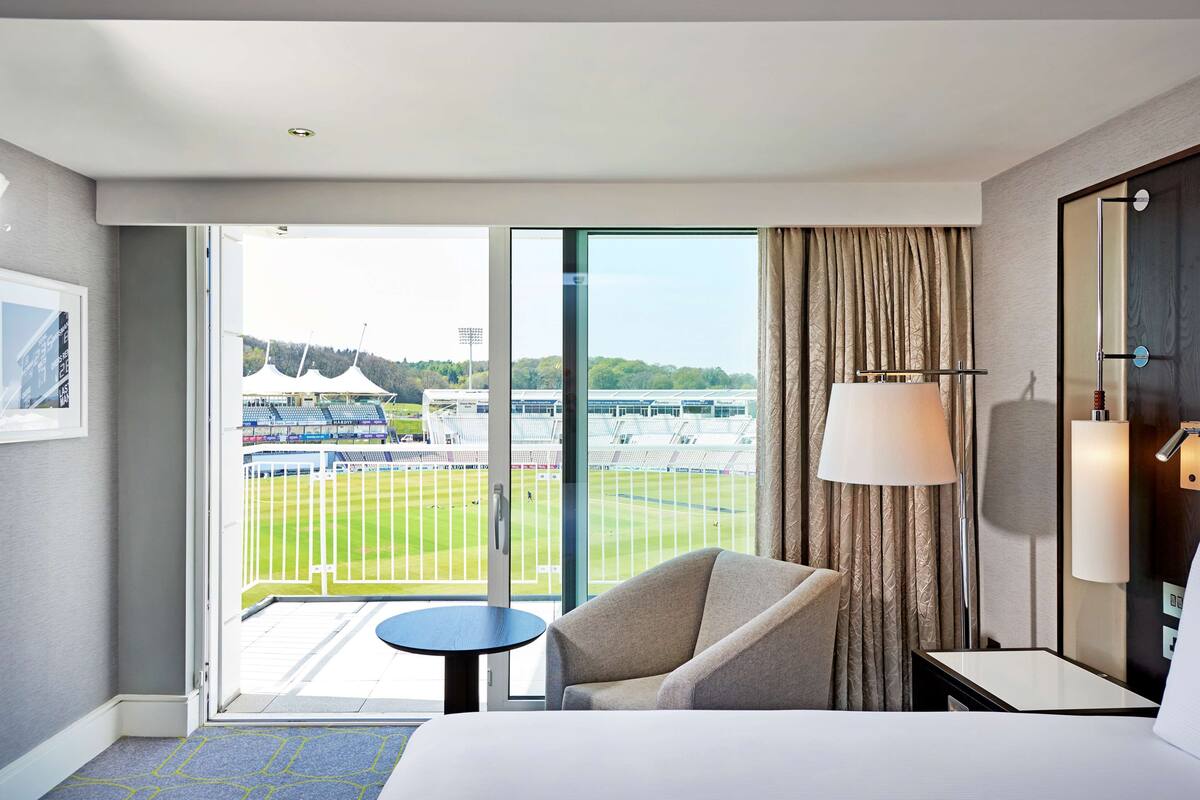 Een hotelkamer met een bed, een stoel, een tafeltje, een lamp en uitzicht op een stadion door een groot raam.