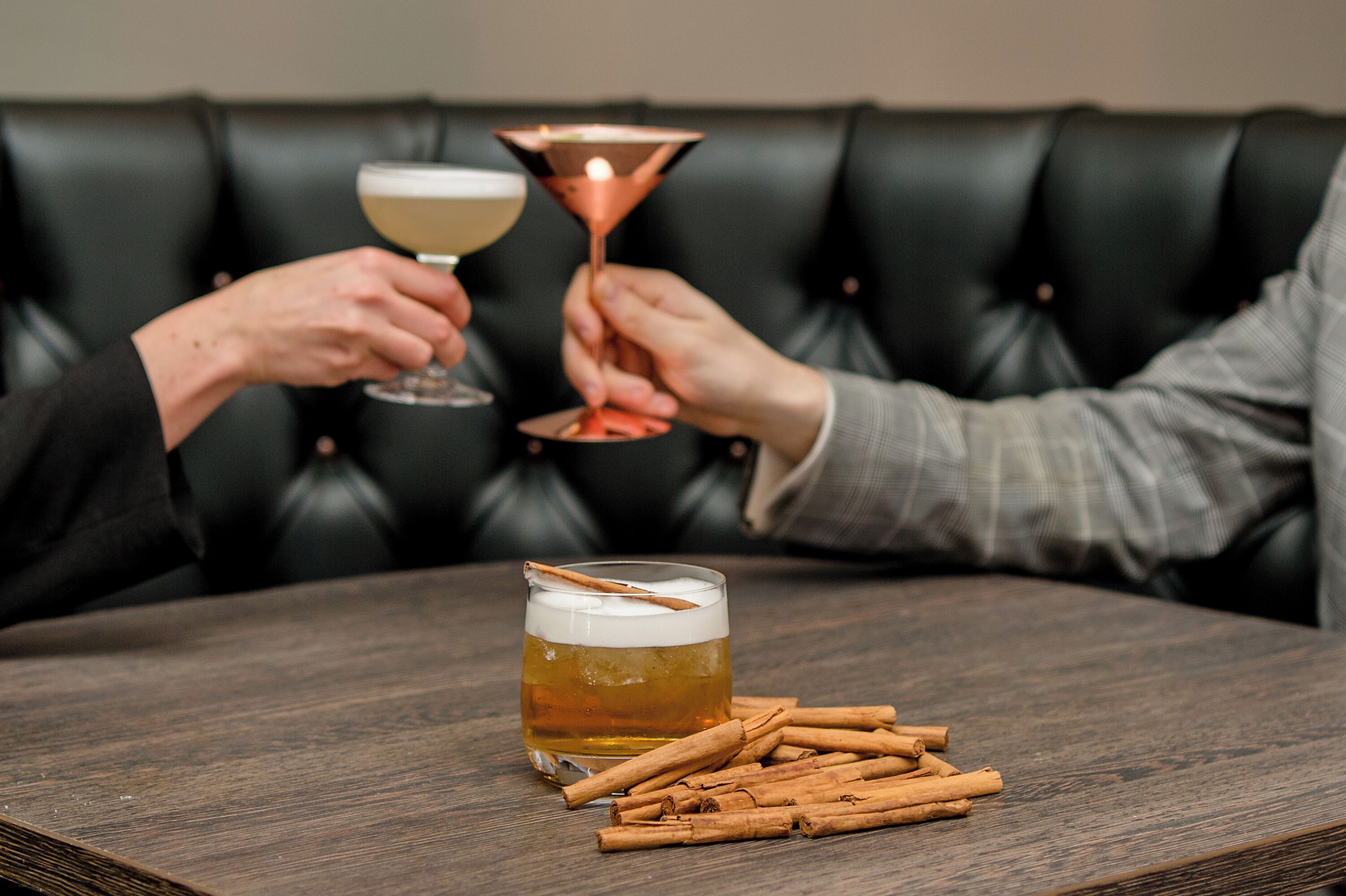 Zwei Personen stoßen mit Martiniglasen an, ein Bierglas mit Zimtstange, und Zimtstangen auf einem Holztisch.