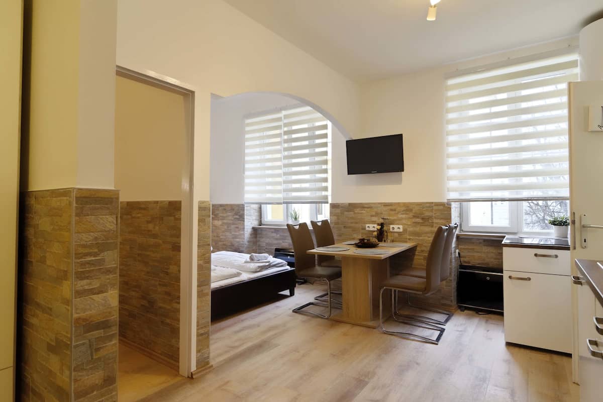Habitación de hotel pequeña con una cama, una mesa de comedor con sillas, un televisor montado en la pared y una ventana con persianas.