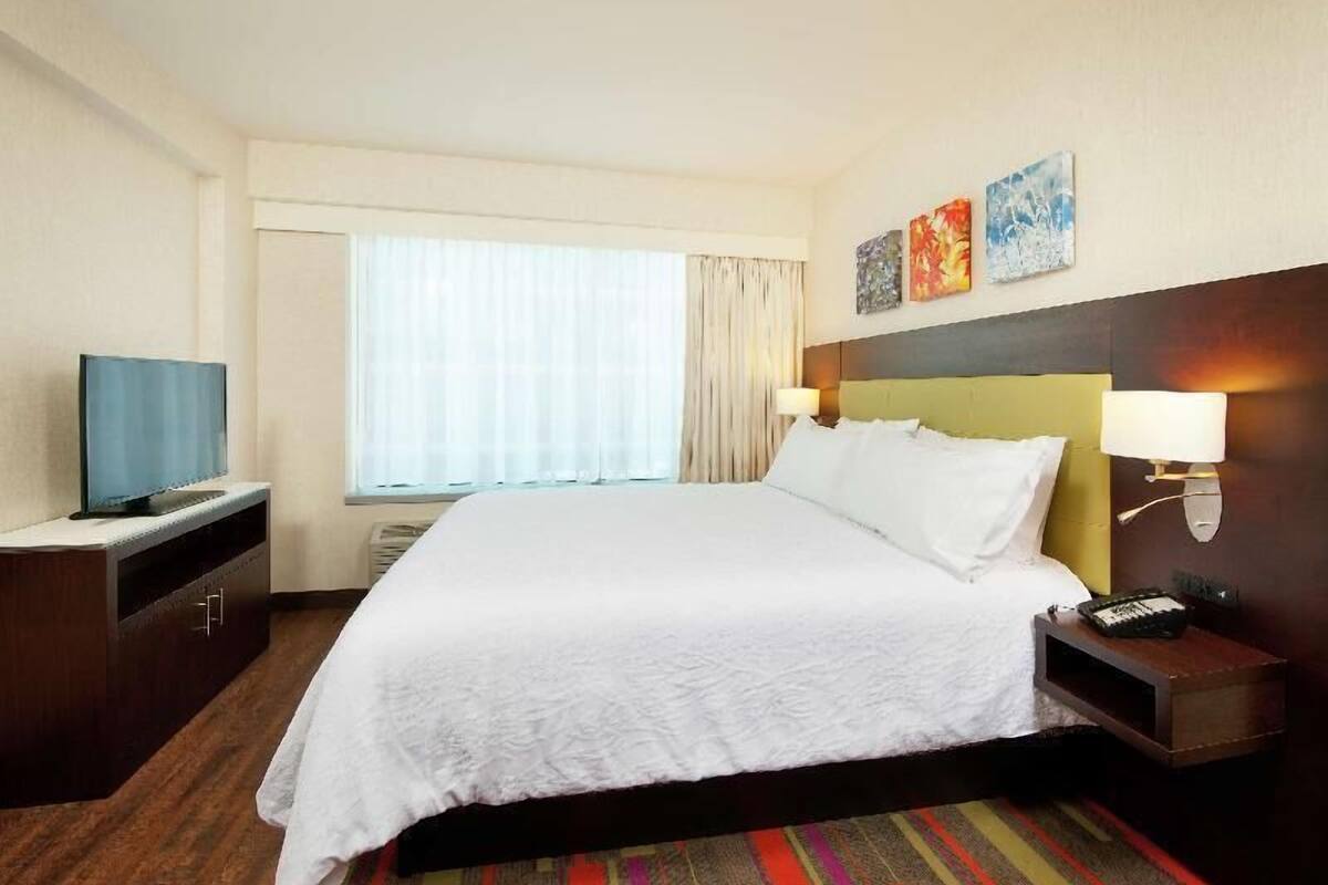 A hotel room with a large bed, a television on a desk, a window with curtains, and artwork on the wall.