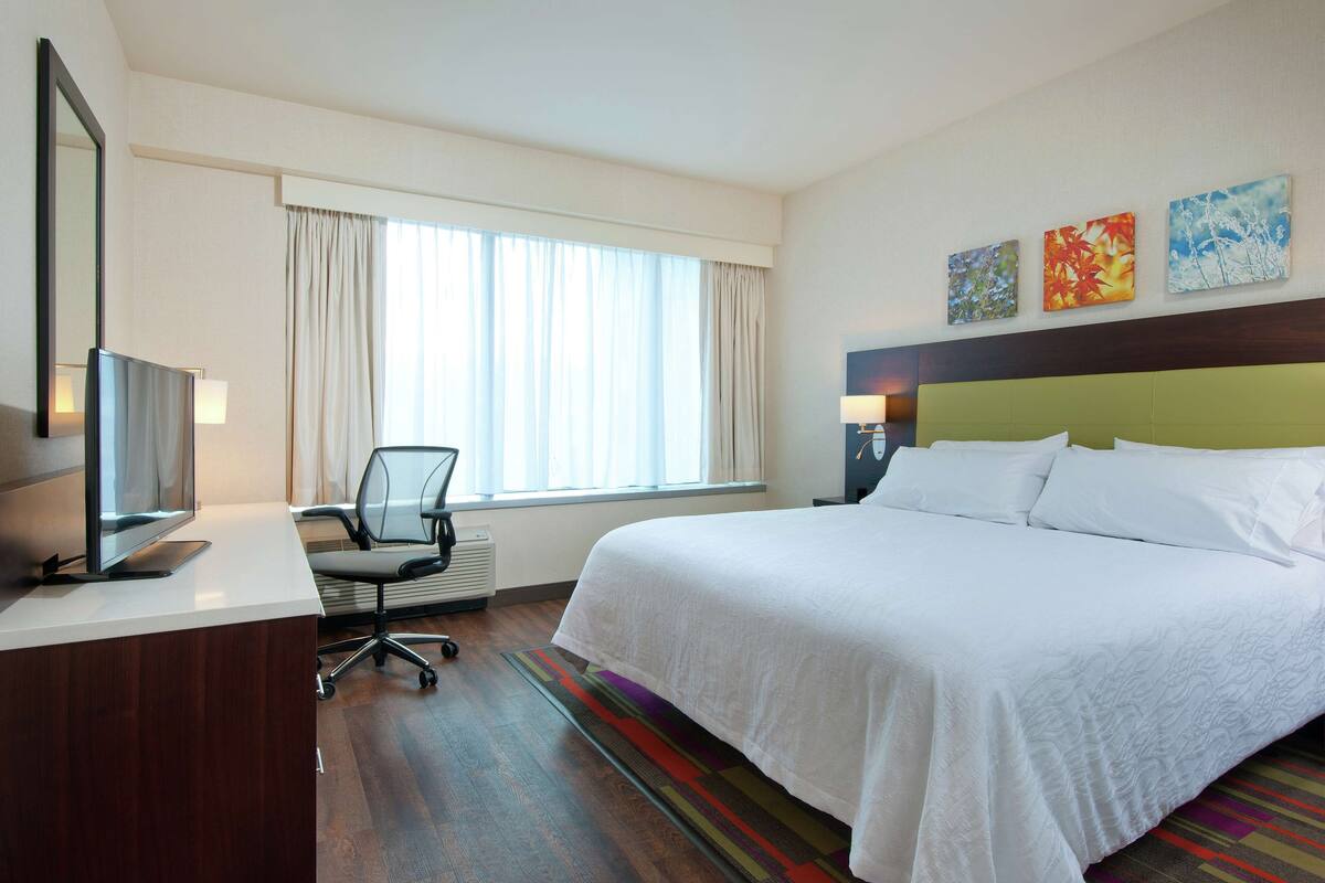 A hotel room with a large bed, a desk with a computer, a chair, a TV, and a window with curtains.