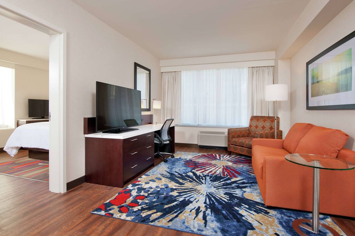 A hotel room with a bed, a desk with a TV, an orange sofa, a glass coffee table, and a colorful rug.