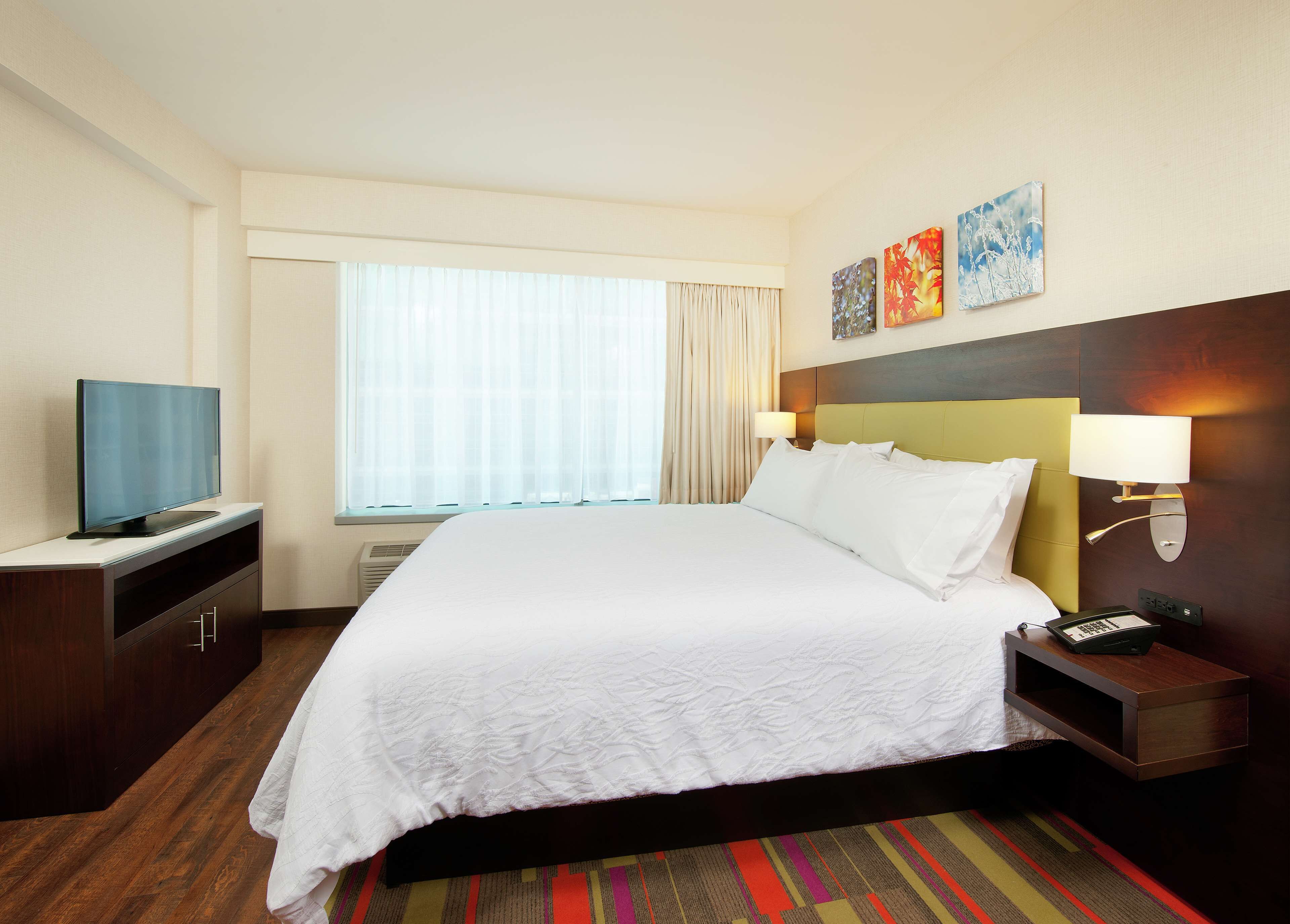 Une chambre d’hôtel avec un grand lit, un téléviseur posé sur un meuble, une table de chevet avec une lampe et des œuvres d’art accrochées au mur.