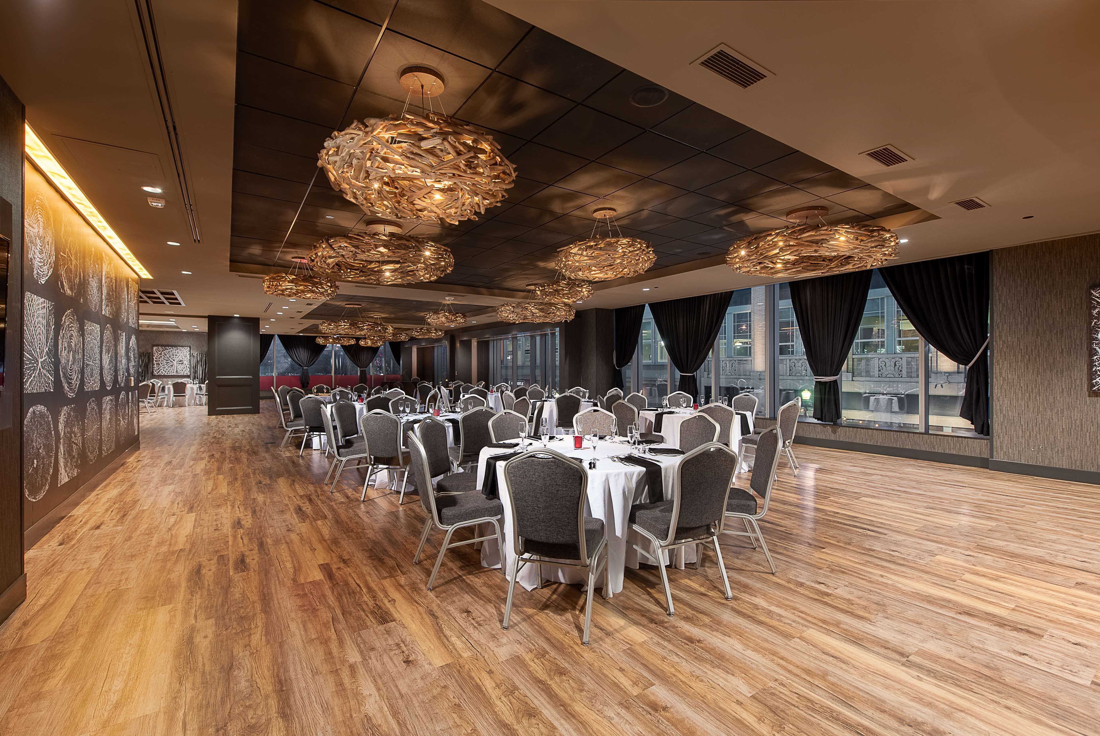 Une salle de banquet spacieuse, dotée de nombreuses tables rondes et de chaises, de grandes fenêtres avec des rideaux et de luminaires décoratifs suspendus au plafond.