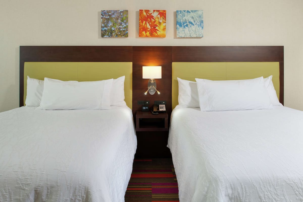 A hotel room with two beds, a nightstand with a lamp, and three wall-mounted artworks.