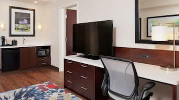 Quarto de hotel com televisão de tela plana, uma escrivaninha com cadeira, pequena cozinha, micro-ondas, geladeira, tapete estampado e quadro na parede.