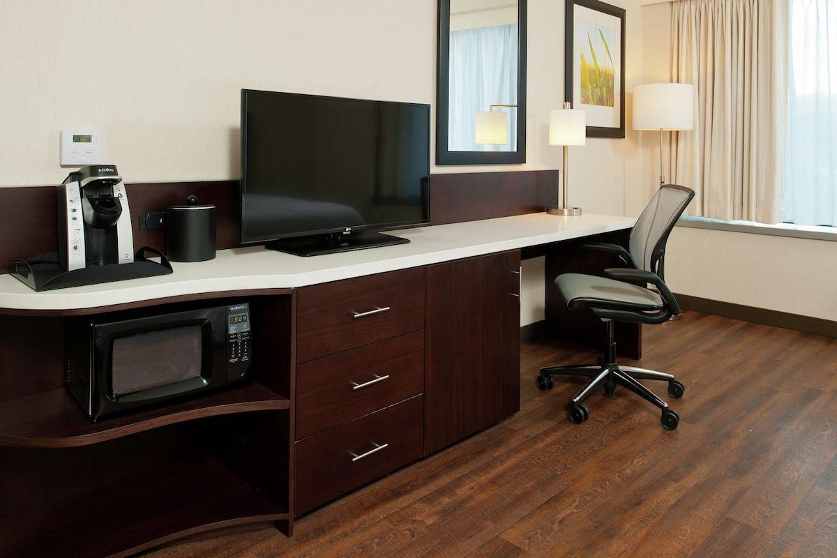 A hotel room with a desk, a flat-screen TV, a microwave, a coffee maker, a lamp, and a chair.