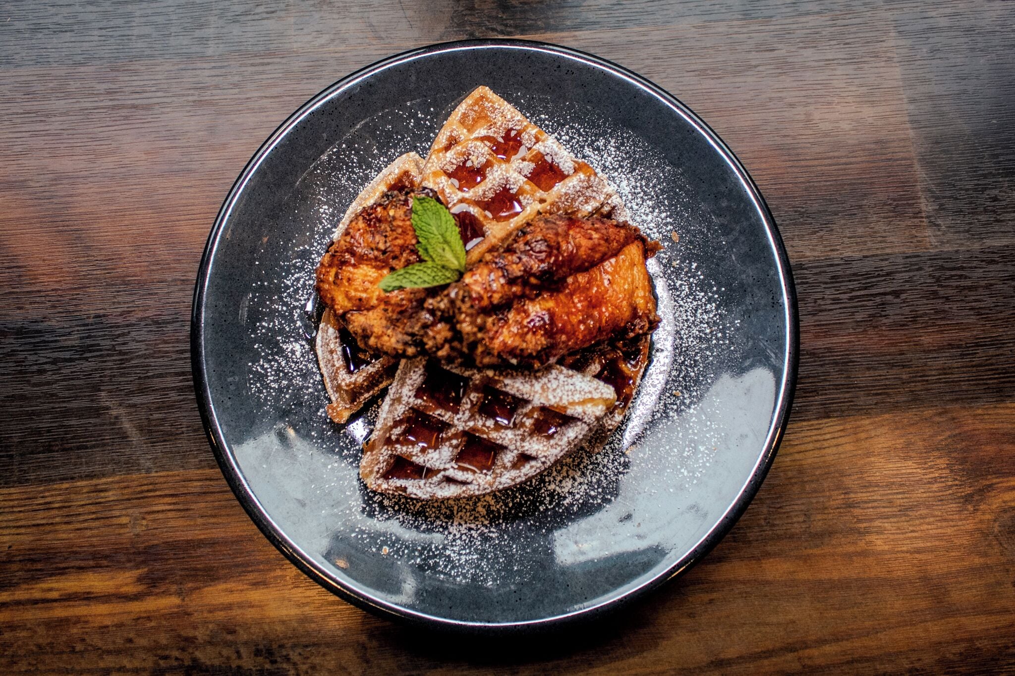 A plate with waffles, syrup, and a mint leaf on a dark plate.