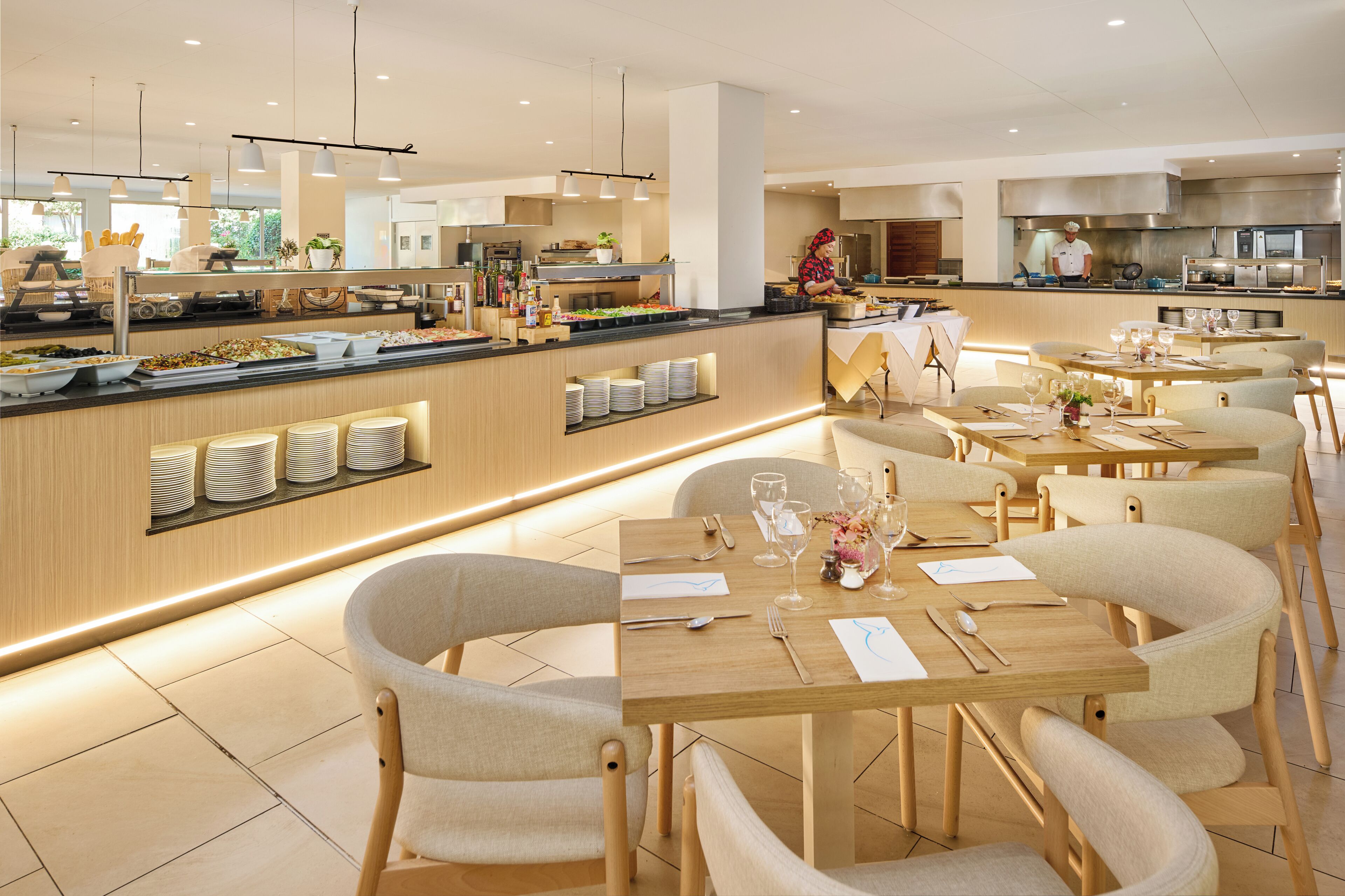 Un espace de restauration moderne avec des tables et des chaises en bois, un buffet proposant divers plats, et une cuisine en arrière-plan.