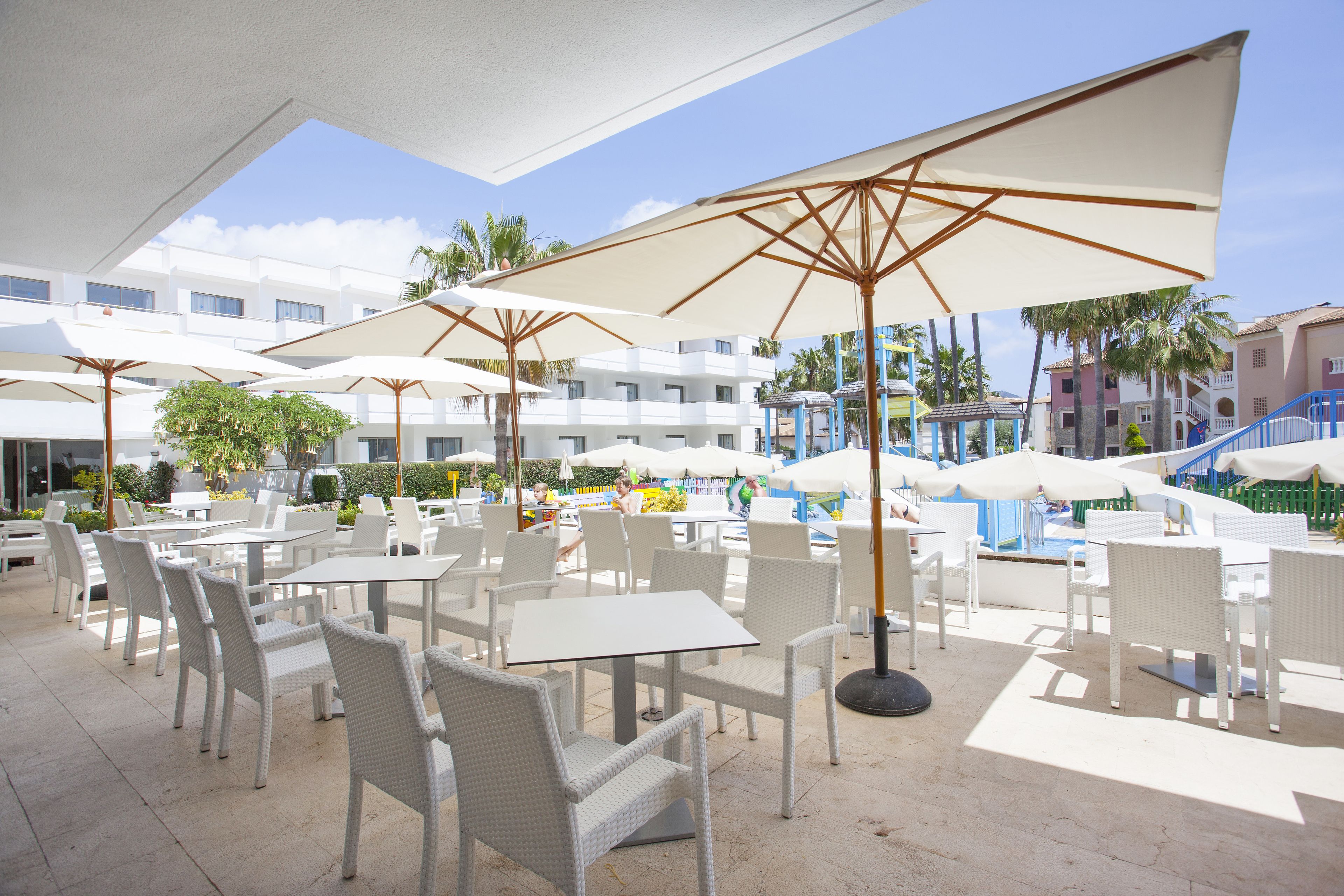 An outdoor patio area with white wicker chairs and tables, large umbrellas, and a pool in the background.