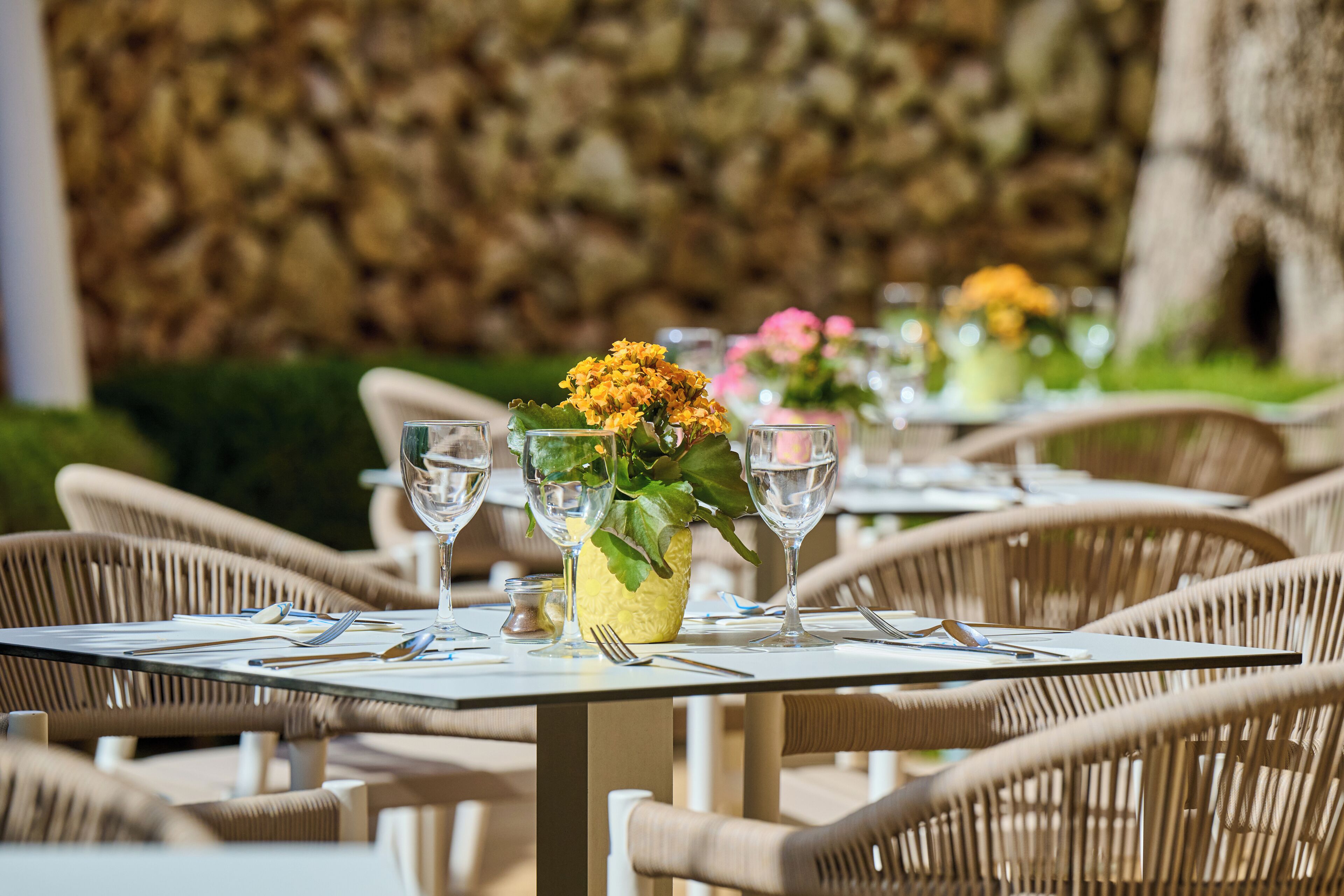 A dining table set with clear glassware and a vase with yellow flowers, surrounded by wicker chairs.