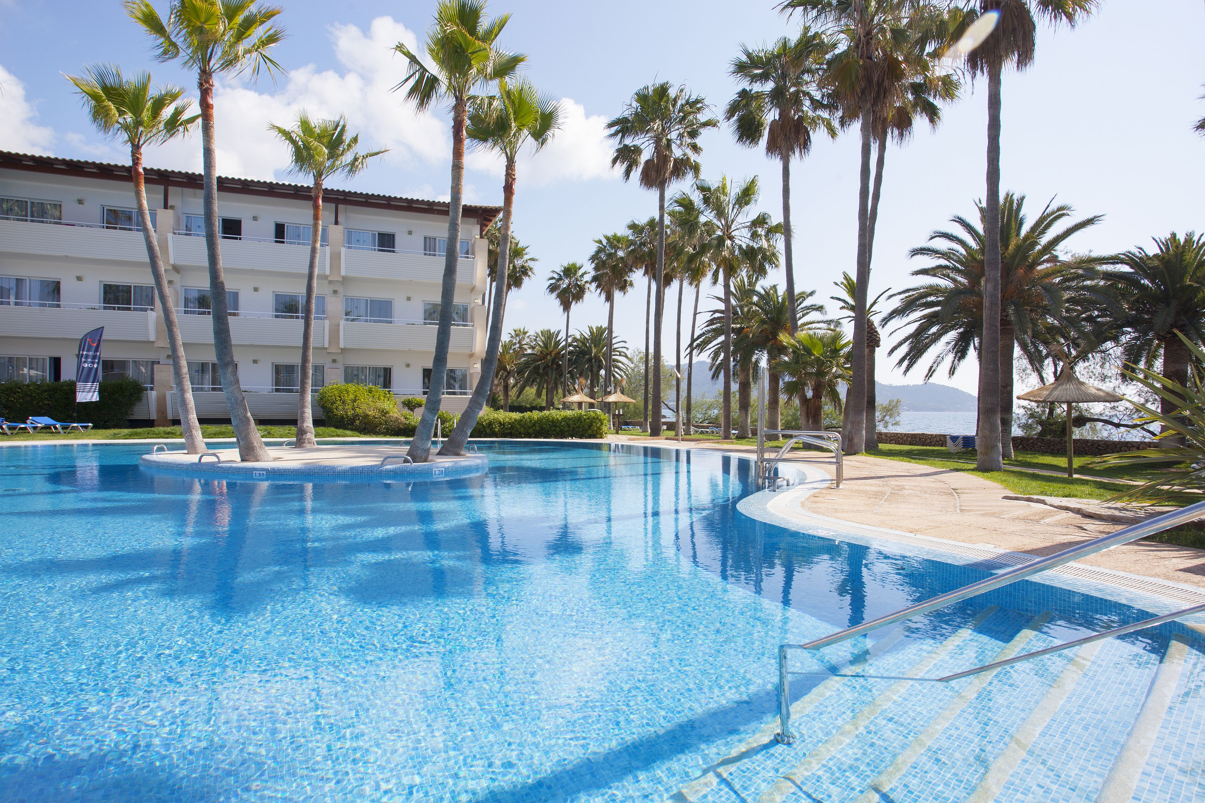 A swimming pool with palm trees, a building with multiple balconies, and a clear sky.