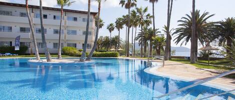 A swimming pool with palm trees, a building with multiple balconies, and a clear sky.