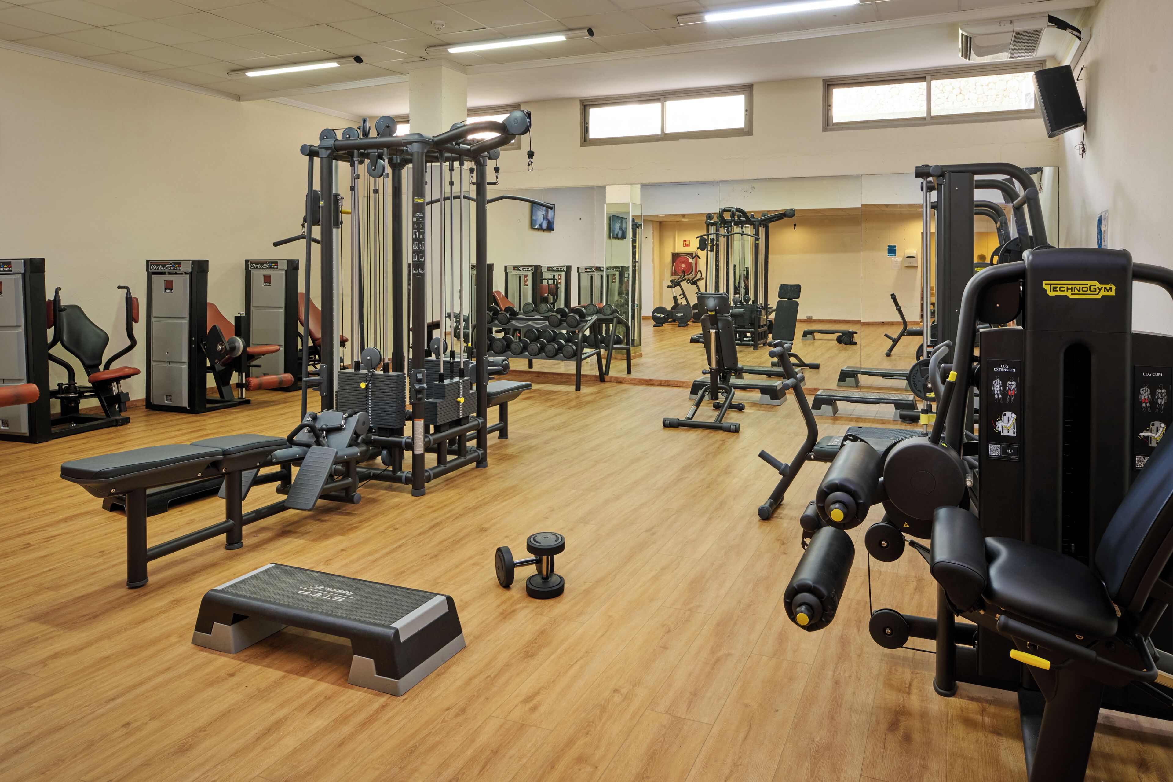 Een sportschool met diverse fitnessapparaten, waaronder krachttoestellen, losse gewichten en een bank.