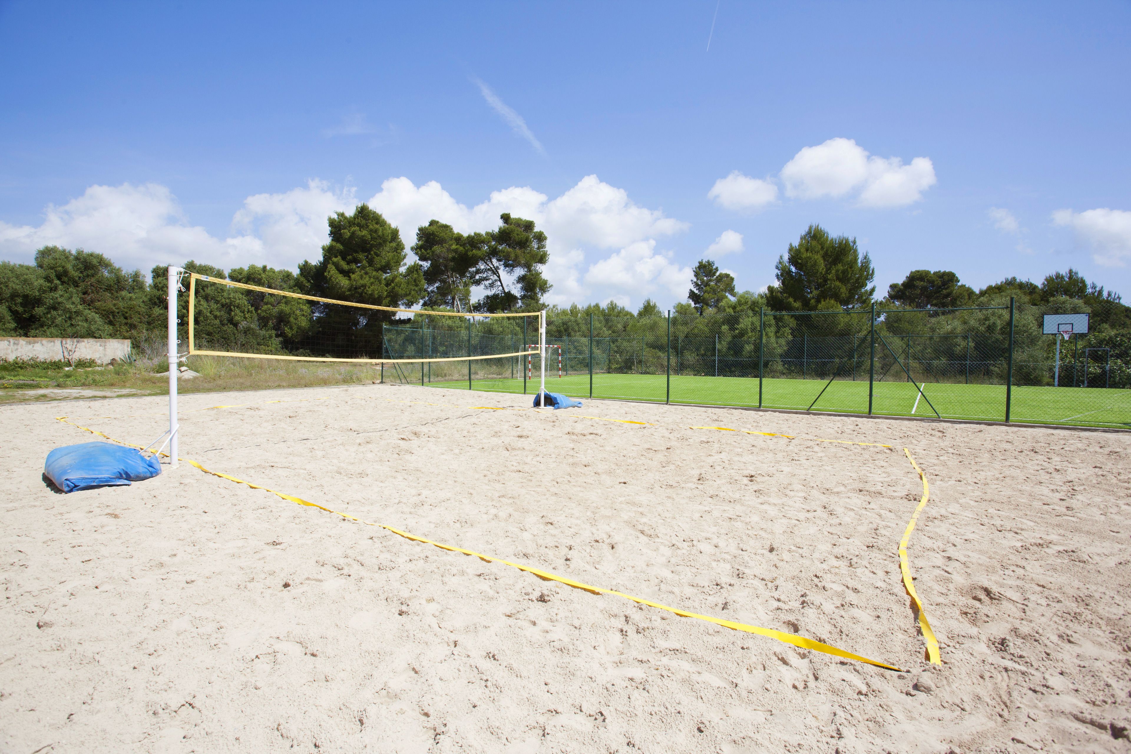 A sandy outdoor sports court with a volleyball net, surrounded by a fence and trees.