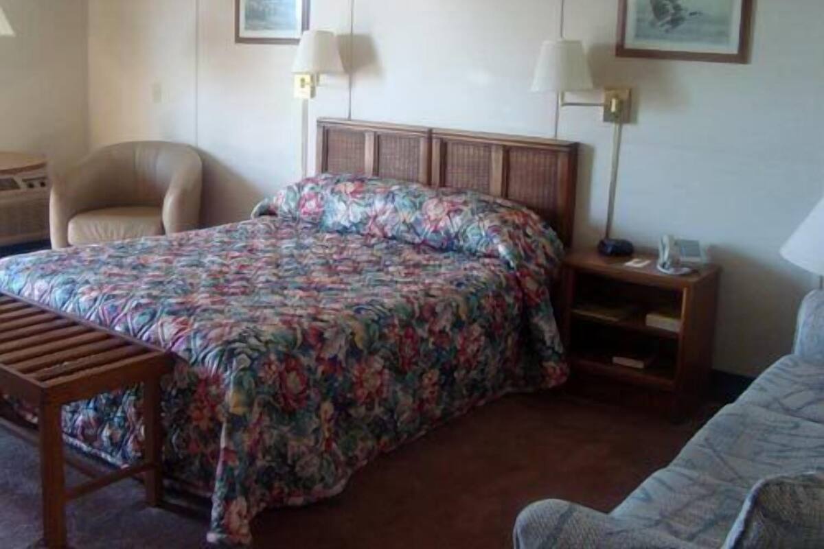 Quarto de hotel com cama, uma cadeira, mesa de cabeceira e abajur.