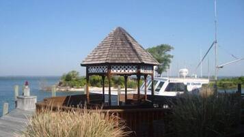 Un kiosque doté d’une structure en bois et d’un motif en treillis, situé près d’une étendue d’eau, avec un bateau amarré au loin.
