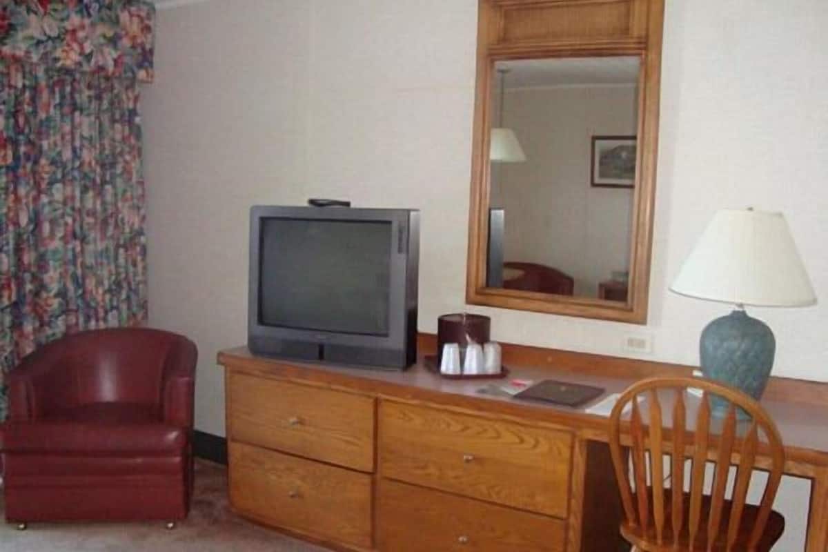 Quarto com cômoda de madeira, televisão, espelho, cadeira vermelha e escrivaninha de madeira com abajur.