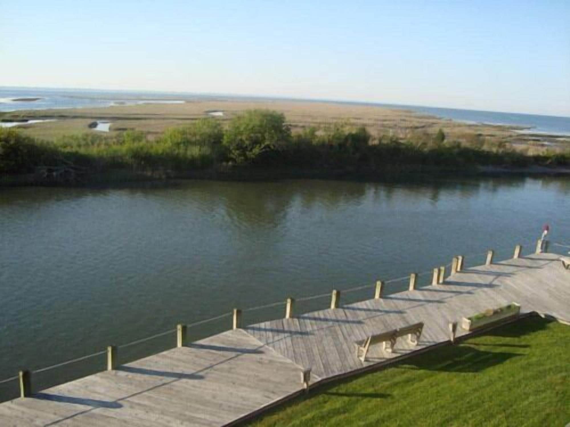 Una passerella in legno che si estende sull'acqua, con una zona erbosa e una costa in lontananza.