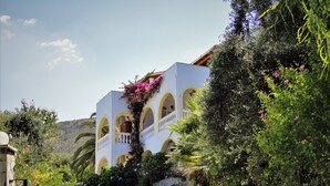 A white building with arched windows and balconies, surrounded by lush greenery and a gravel path leading to the entrance. - Dionysos Apartments (Corfu)