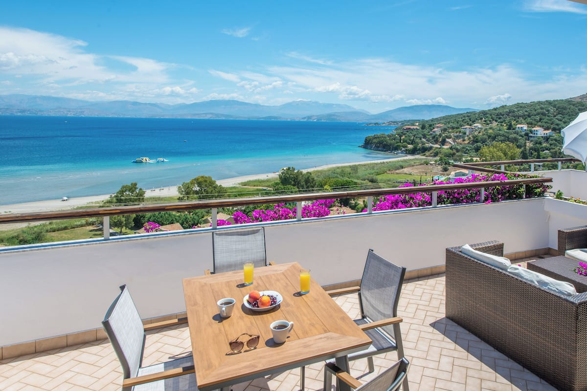 Un balcón con una mesa de madera, sillas y vistas al mar y las montañas.