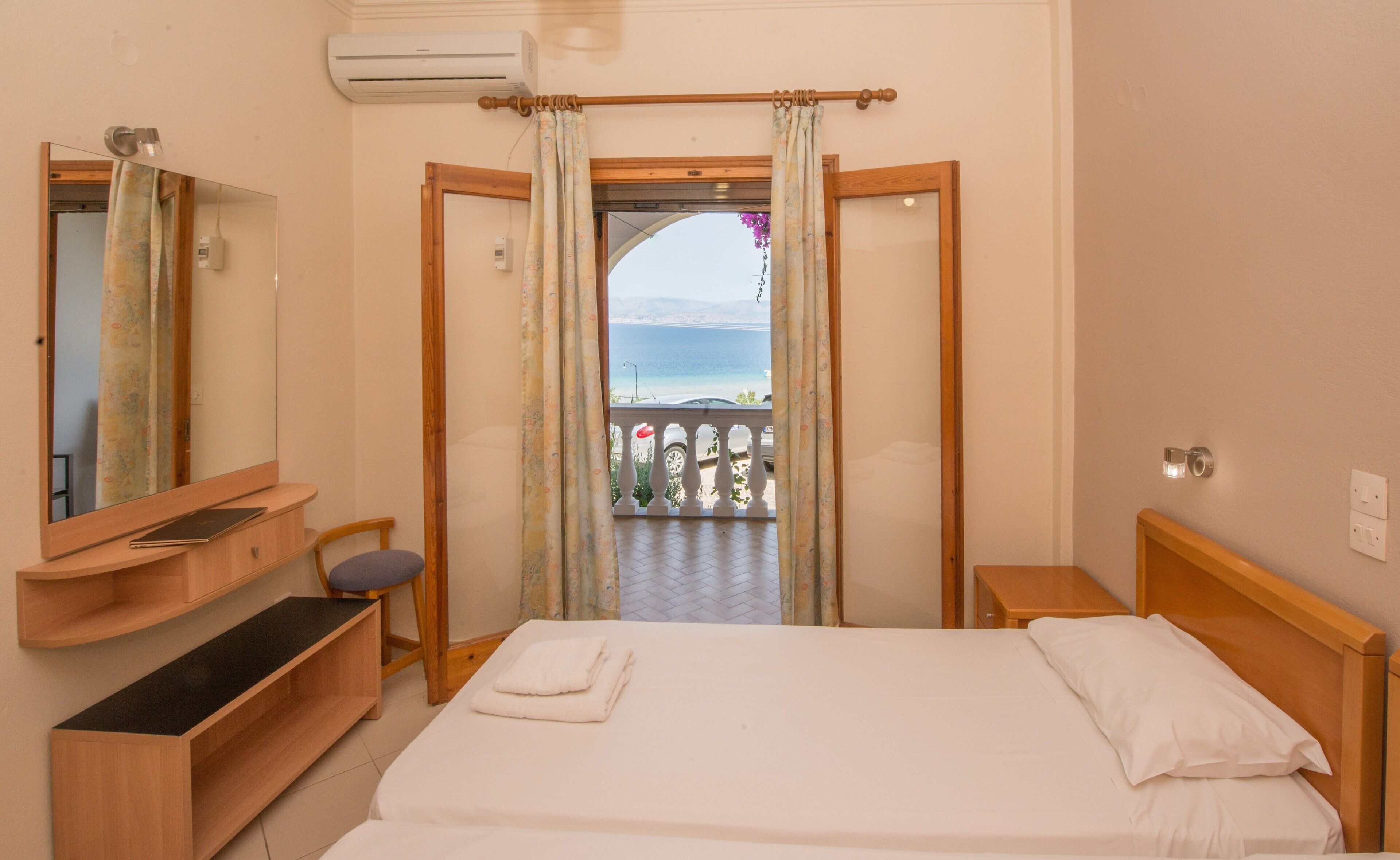A hotel room with a bed, a wooden nightstand, a bedside table, a mirror, a bathroom mirror, a window with curtains, and an air conditioning unit.