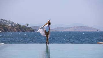 A person standing on a pool deck with a view of the sea and hills in the background.