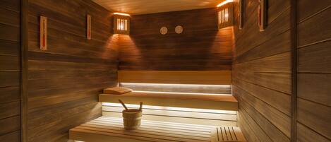A sauna with wooden walls and benches, a bucket, and two wall-mounted clocks.