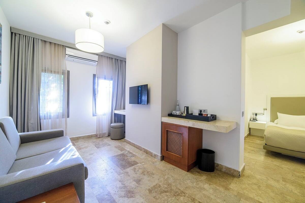 A hotel room with a sofa, a small table with a lamp, a TV mounted on the wall, and a bed with a headboard.