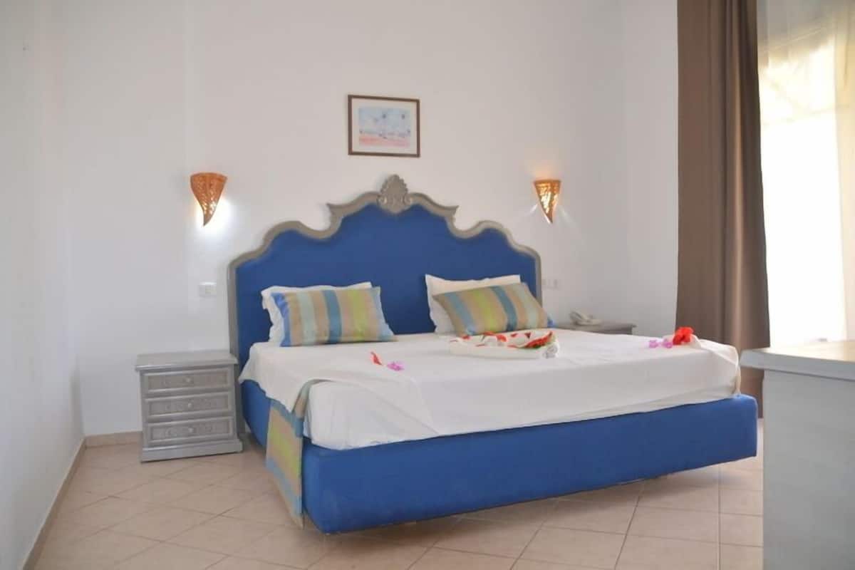 A bedroom with a blue bed, two pillows, a nightstand, and two wall-mounted lights.