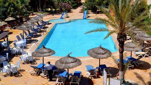 A pool area with lounge chairs and umbrellas, surrounded by palm trees and a clear blue sky. - Diar Yassine (Djerba Midun)