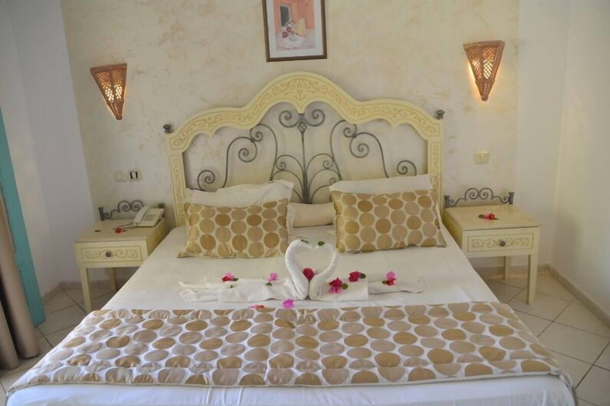 A neatly made bed with a patterned bedspread, two matching pillows, and a decorative swan design on the bed. There are two bedside tables with lamps and a small table with a flower arrangement.