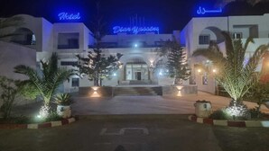 A hotel with a modern facade, illuminated signage, and a well-lit entrance area with palm trees. - Diar Yassine (Djerba Midun)