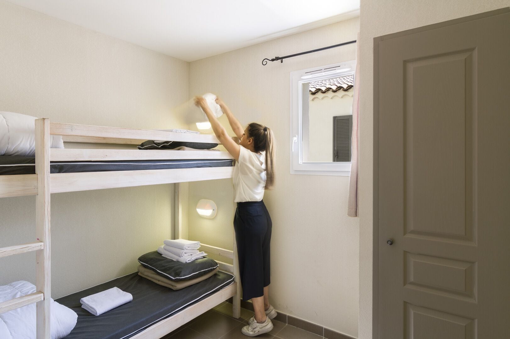 A bunk bed room with a person adjusting the top bunk. There is a window with a view of a building with a tiled roof.