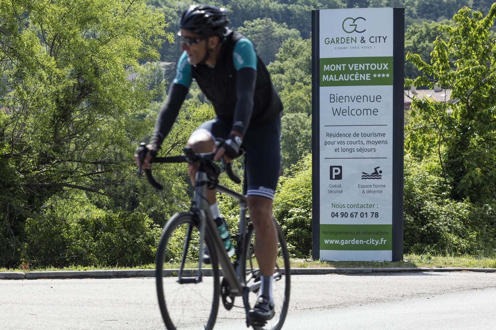 Ciclista en una carretera con un cartel de Garden & City que indica la bienvenida al Mont Ventoux y Malaucene.