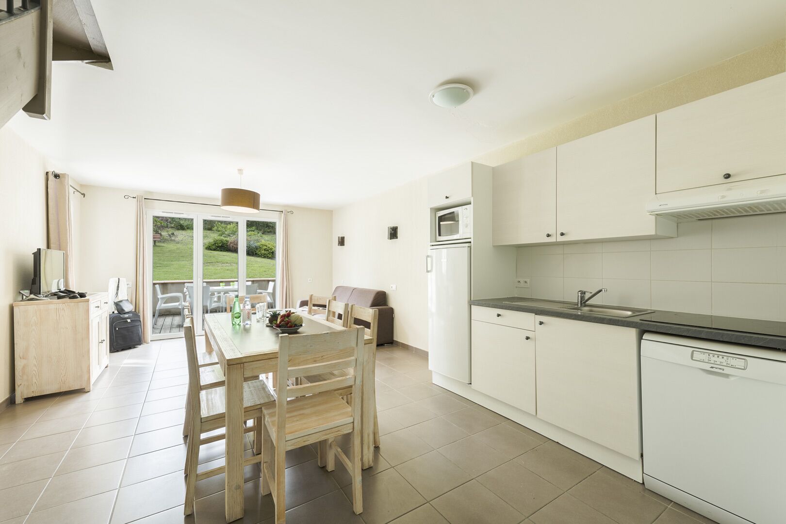 Una cocina moderna con armarios blancos, un comedor con sillas de madera y vistas a un jardín a través de una puerta corrediza de vidrio.